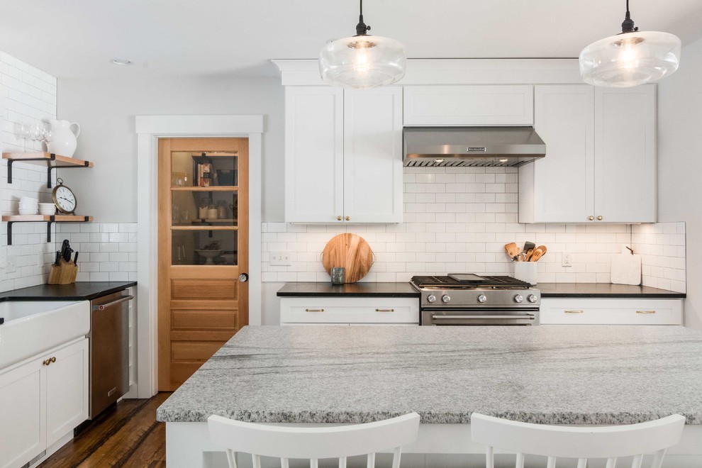 Design ideas for a mid-sized country l-shaped eat-in kitchen in Portland Maine with a farmhouse sink, shaker cabinets, white cabinets, soapstone benchtops, white splashback, subway tile splashback, stainless steel appliances, medium hardwood floors, with island, brown floor and black benchtop.