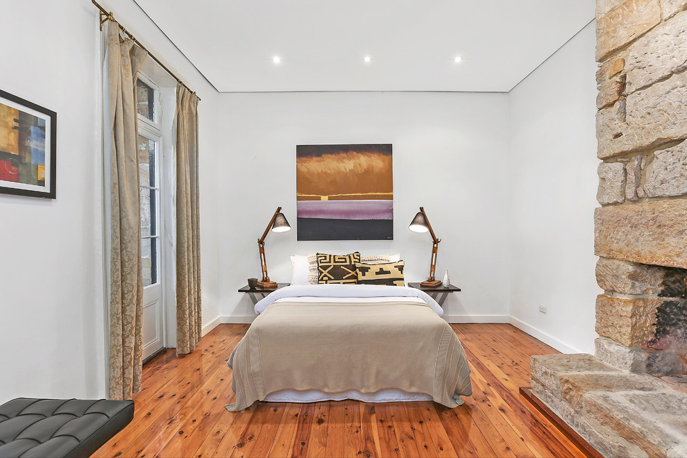 This is an example of a transitional bedroom in Sydney with white walls, medium hardwood floors, a standard fireplace, a stone fireplace surround and brown floor.