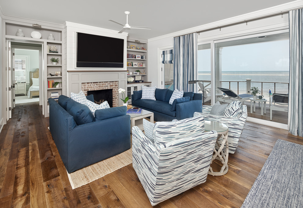 Photo of a large beach style open concept family room in Charleston with white walls, medium hardwood floors, a standard fireplace, a brick fireplace surround, a wall-mounted tv and brown floor.