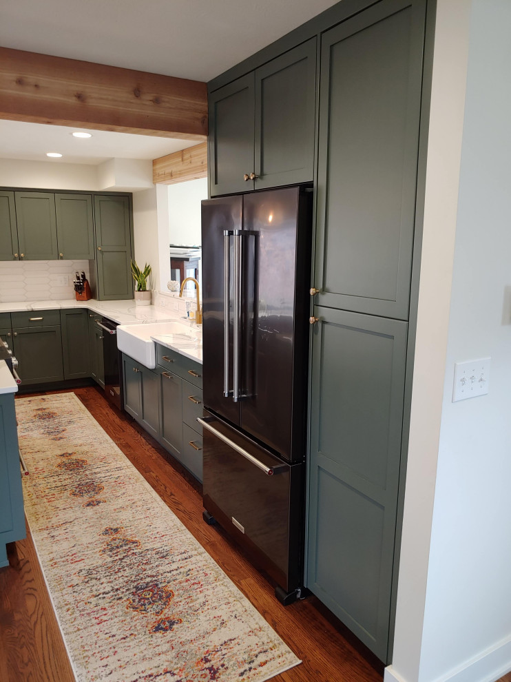 Example of a mid-sized transitional l-shaped dark wood floor eat-in kitchen design with a farmhouse sink, recessed-panel cabinets, green cabinets, quartz countertops, white backsplash, black appliances, an island and white countertops