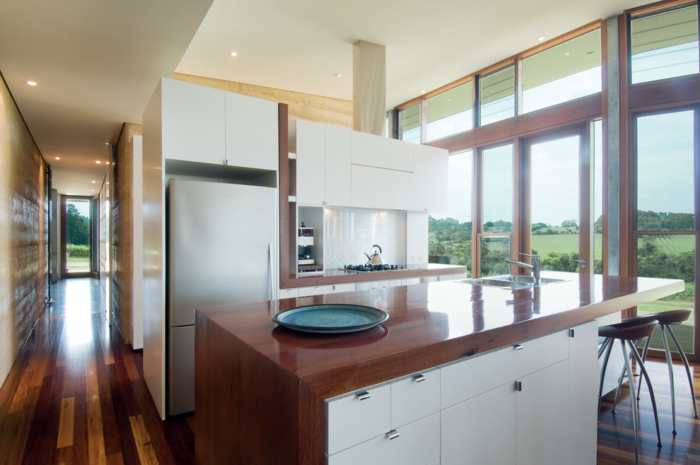 Inspiration for an expansive modern galley open plan kitchen in Melbourne with a drop-in sink, flat-panel cabinets, stainless steel appliances, wood benchtops, white cabinets, white splashback, glass sheet splashback, dark hardwood floors and with island.