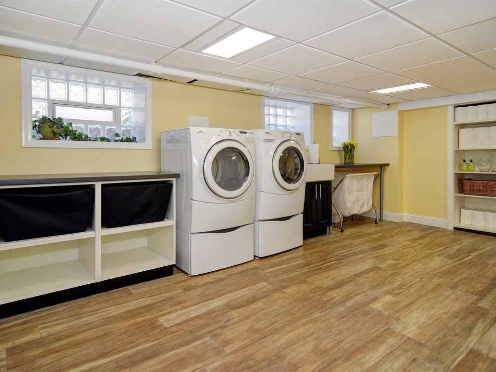 This is an example of an expansive transitional single-wall dedicated laundry room in Newark with a farmhouse sink, shaker cabinets, black cabinets, laminate benchtops, yellow walls, porcelain floors and a side-by-side washer and dryer.
