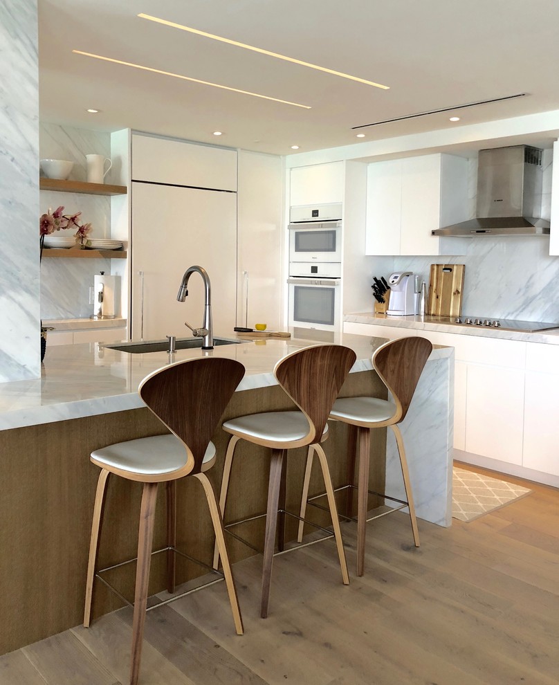 Inspiration for a mid-sized contemporary galley kitchen in Miami with an undermount sink, flat-panel cabinets, white cabinets, marble benchtops, grey splashback, marble splashback, light hardwood floors, beige floor, white appliances, a peninsula and grey benchtop.