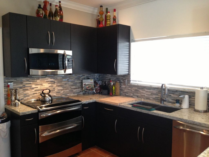 Example of a mid-sized u-shaped terra-cotta tile eat-in kitchen design in Miami with a double-bowl sink, flat-panel cabinets, black cabinets, stainless steel countertops, gray backsplash, matchstick tile backsplash, stainless steel appliances and no island