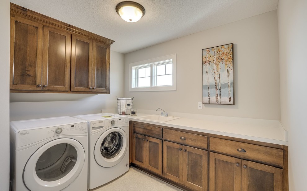 Photo of a mid-sized transitional l-shaped dedicated laundry room in Minneapolis with a drop-in sink, beaded inset cabinets, medium wood cabinets, laminate benchtops, grey walls, linoleum floors, a side-by-side washer and dryer and white floor.
