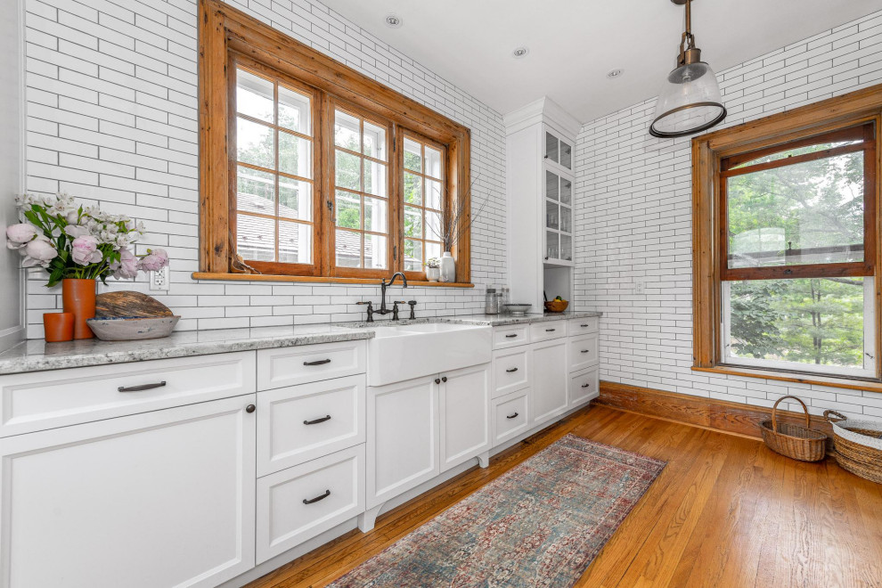Small transitional galley light wood floor and brown floor enclosed kitchen photo in Ottawa with a farmhouse sink, shaker cabinets, white cabinets, granite countertops, white backsplash, subway tile backsplash, stainless steel appliances, an island and gray countertops