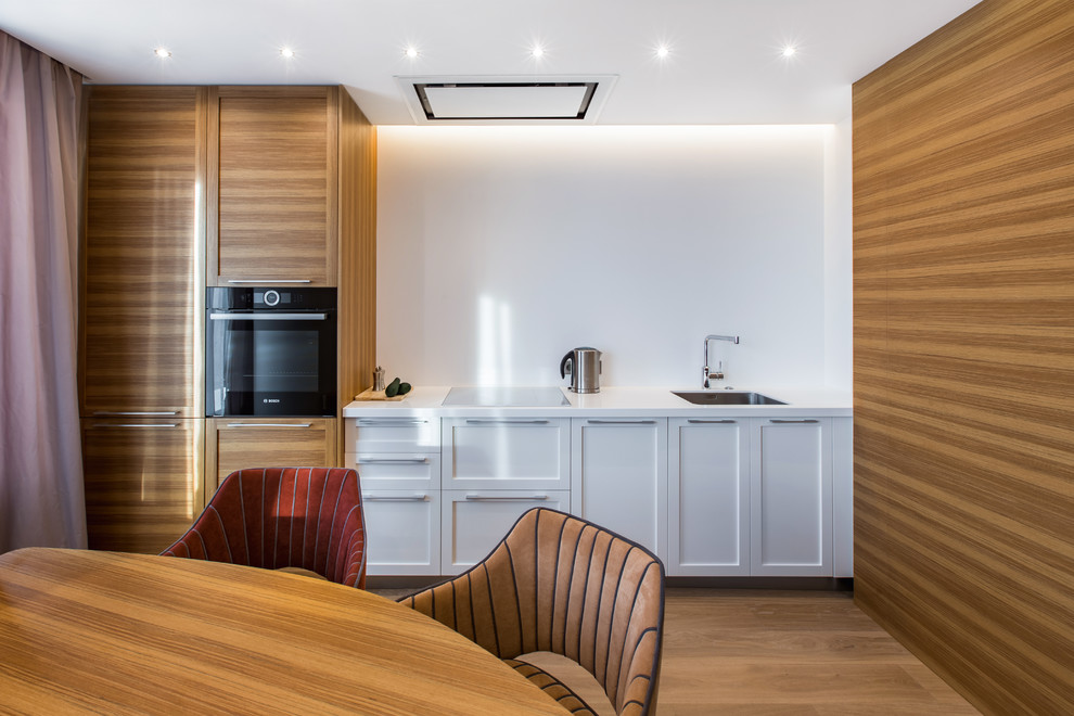 This is an example of a contemporary single-wall eat-in kitchen in Los Angeles with an undermount sink, white cabinets, white splashback, black appliances, medium hardwood floors, no island, brown floor, white benchtop and shaker cabinets.