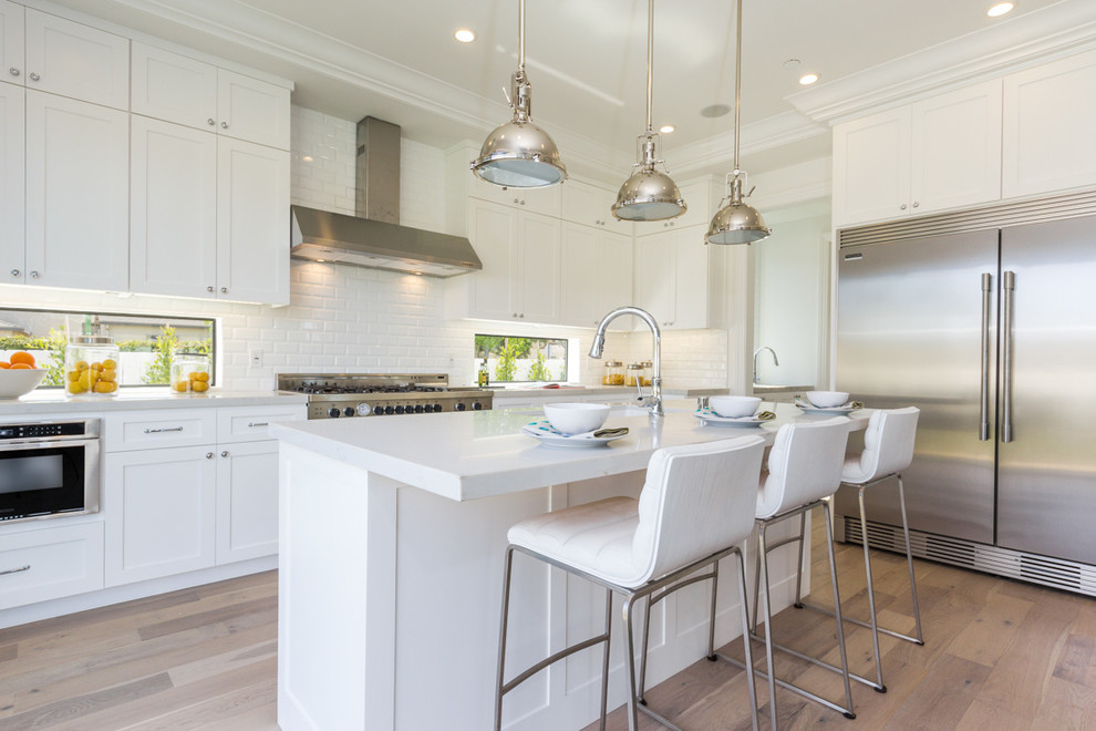 Design ideas for a transitional l-shaped kitchen in Los Angeles with shaker cabinets, white cabinets, white splashback, subway tile splashback, stainless steel appliances, light hardwood floors, with island and beige floor.