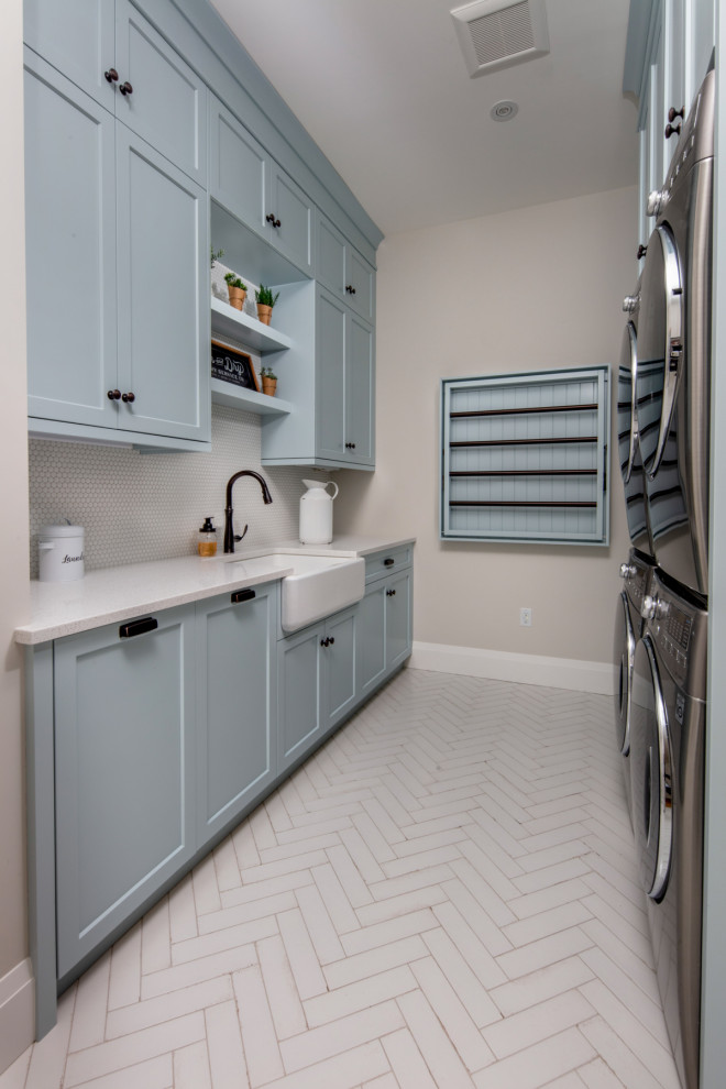 Mid-sized country galley dedicated laundry room in Toronto with a farmhouse sink, shaker cabinets, blue cabinets, quartz benchtops, grey splashback, ceramic splashback, beige walls, a stacked washer and dryer, white floor and white benchtop.