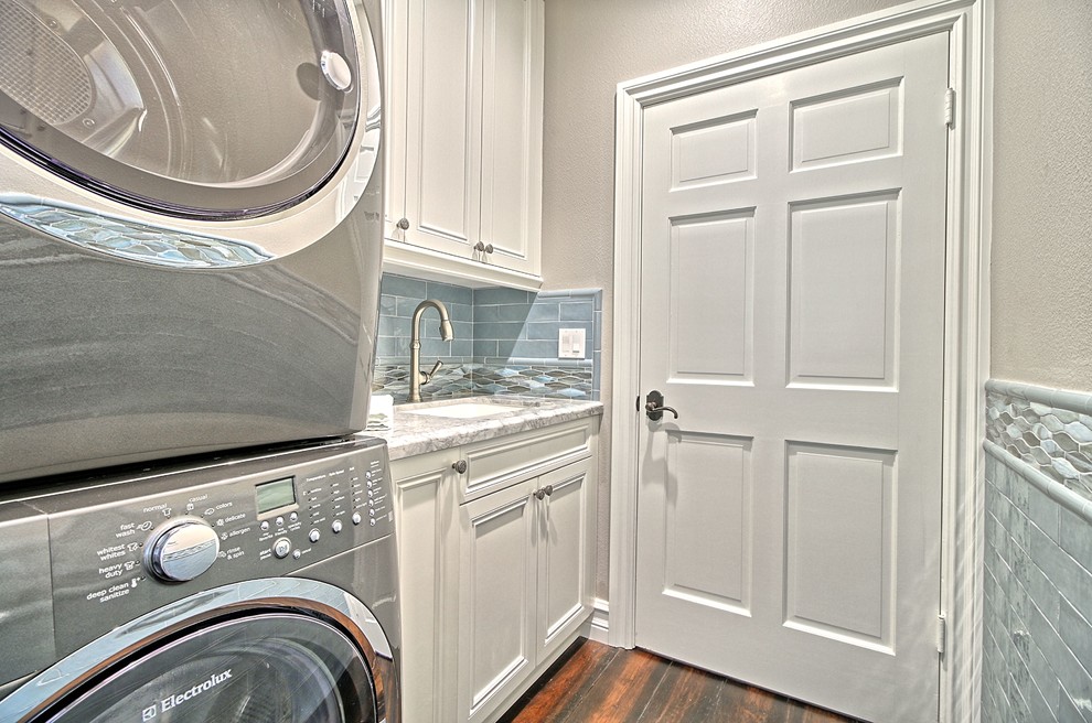 Design ideas for a mid-sized transitional single-wall dedicated laundry room in Los Angeles with an undermount sink, recessed-panel cabinets, white cabinets, quartz benchtops, grey walls, medium hardwood floors, a stacked washer and dryer and brown floor.