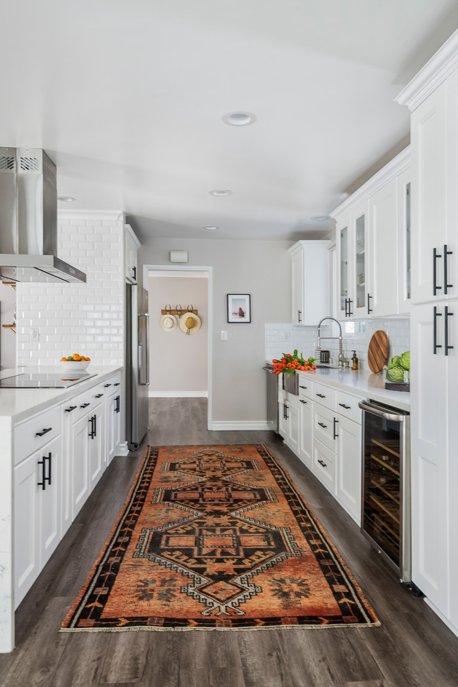 Inspiration for a transitional galley kitchen in Los Angeles with a farmhouse sink, shaker cabinets, white cabinets, subway tile splashback, stainless steel appliances, medium hardwood floors, a peninsula, brown floor and white benchtop.