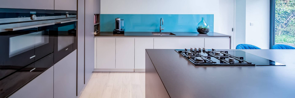 Photo of a large contemporary l-shaped open plan kitchen in London with a single-bowl sink, flat-panel cabinets, grey cabinets, quartz worktops, blue splashback, black appliances, light hardwood flooring, an island, beige floors and grey worktops.