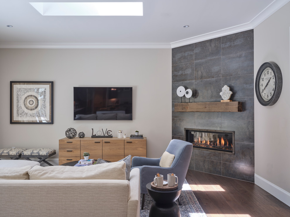 Modelo de salón cerrado de estilo de casa de campo de tamaño medio con paredes beige, suelo de madera en tonos medios, chimenea de esquina, marco de chimenea de baldosas y/o azulejos, televisor colgado en la pared y suelo marrón