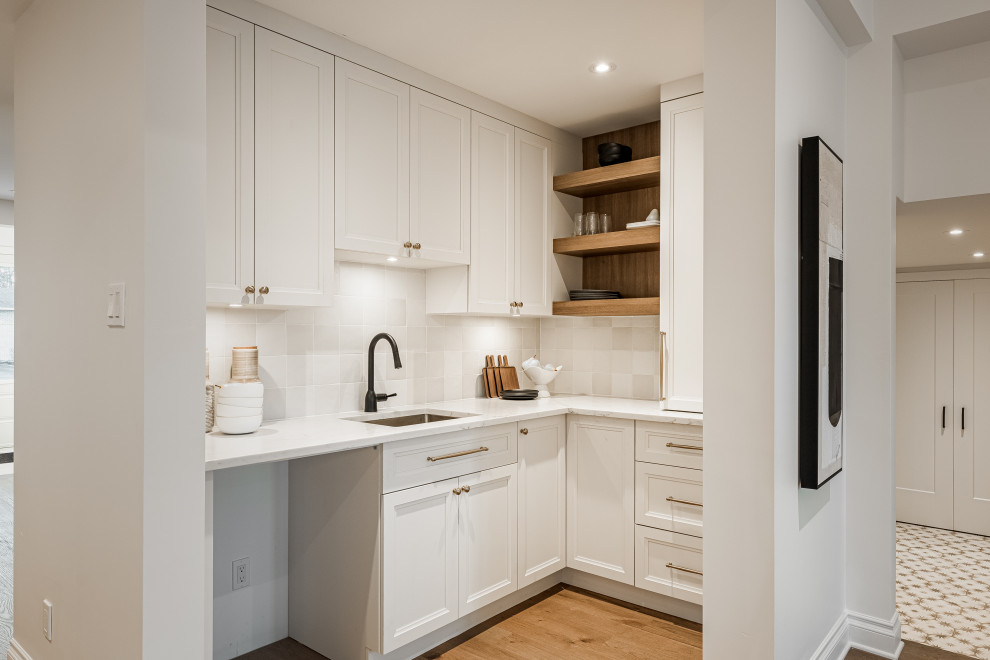 Photo of a small modern l-shaped wet bar in Montreal with an undermount sink, recessed-panel cabinets, white cabinets, quartz benchtops, white splashback, ceramic splashback, light hardwood floors, beige floor and white benchtop.