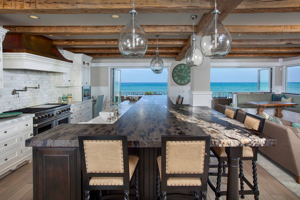 This is an example of a beach style galley open plan kitchen in Orange County with a farmhouse sink, recessed-panel cabinets, white cabinets, white splashback, stainless steel appliances, light hardwood floors and with island.