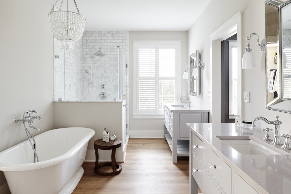 Inspiration for a large country master bathroom in Philadelphia with a freestanding tub, gray tile, marble, grey walls, light hardwood floors, an undermount sink, engineered quartz benchtops, a hinged shower door, grey cabinets, a corner shower, beige floor, grey benchtops and flat-panel cabinets.
