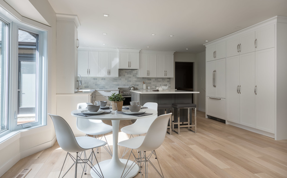 Example of a transitional l-shaped light wood floor eat-in kitchen design with shaker cabinets, white cabinets, solid surface countertops, white backsplash, marble backsplash, stainless steel appliances, an island and white countertops