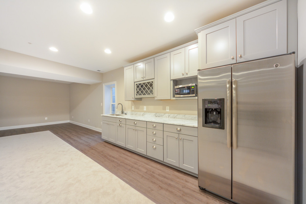 This is an example of a medium sized traditional single-wall wet bar in Baltimore with a submerged sink, shaker cabinets, white cabinets, engineered stone countertops, vinyl flooring, white worktops, feature lighting and brown floors.