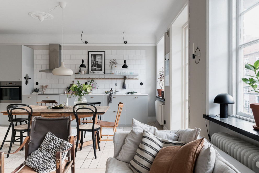 Scandinavian open plan kitchen in Malmo with flat-panel cabinets, grey cabinets, white splashback, ceramic splashback, stainless steel appliances, light hardwood flooring and white worktops.