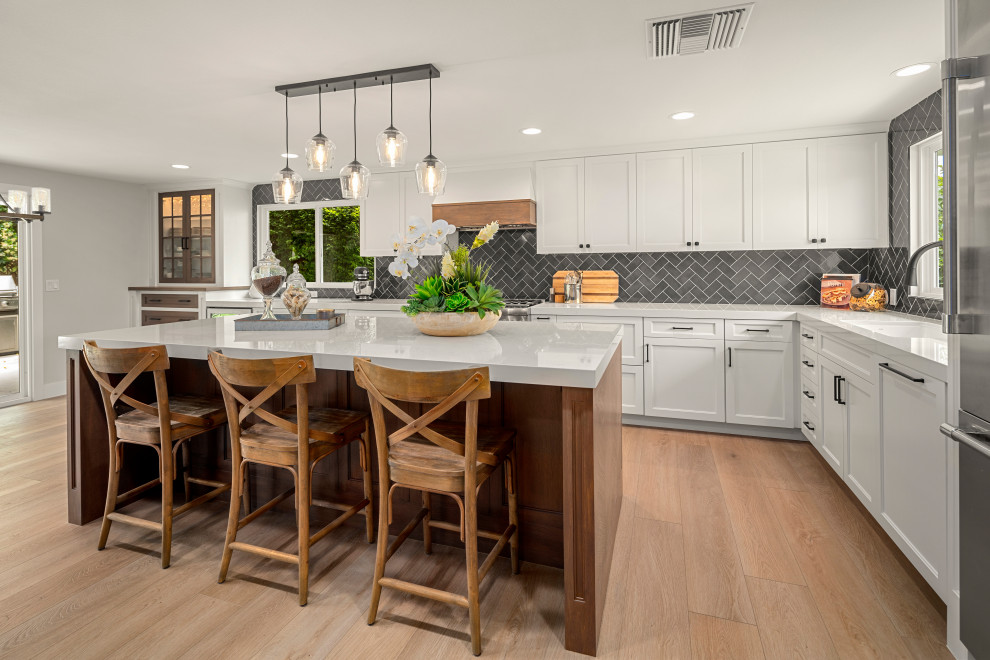 Inspiration for a medium sized farmhouse l-shaped kitchen/diner in Orange County with a belfast sink, shaker cabinets, white cabinets, engineered stone countertops, blue splashback, ceramic splashback, stainless steel appliances, laminate floors and an island.
