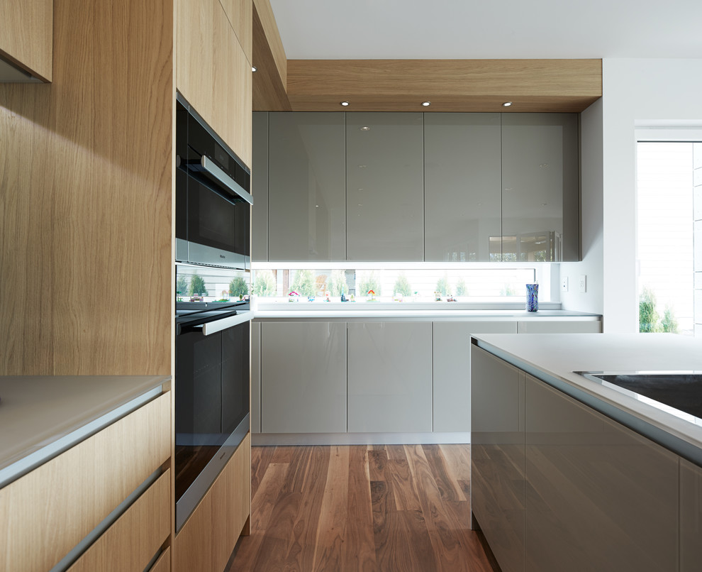 This is an example of a mid-sized contemporary l-shaped open plan kitchen in Vancouver with an undermount sink, flat-panel cabinets, light wood cabinets, glass benchtops, grey splashback, stainless steel appliances, dark hardwood floors and with island.