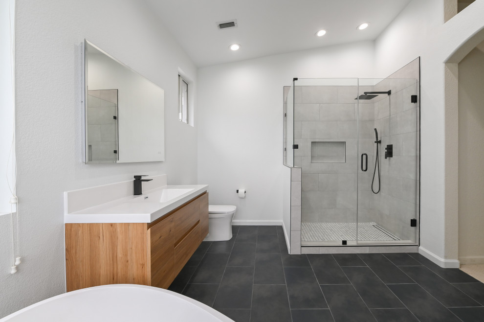 Photo of a mid-sized modern master bathroom in Austin with flat-panel cabinets, medium wood cabinets, a freestanding tub, a double shower, a one-piece toilet, gray tile, ceramic tile, white walls, ceramic floors, a drop-in sink, marble benchtops, black floor, a hinged shower door, white benchtops, a single vanity and a floating vanity.
