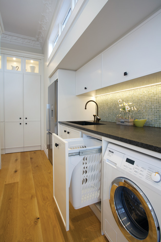 Contemporary galley utility room in Auckland with white cabinets, laminate benchtops, white walls, light hardwood floors, a side-by-side washer and dryer and brown floor.