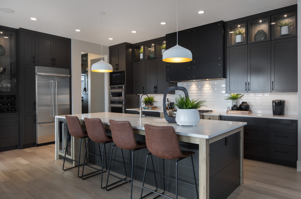 Design ideas for a large country l-shaped eat-in kitchen in Calgary with a farmhouse sink, shaker cabinets, black cabinets, quartz benchtops, white splashback, porcelain splashback, stainless steel appliances, light hardwood floors, with island, brown floor and white benchtop.