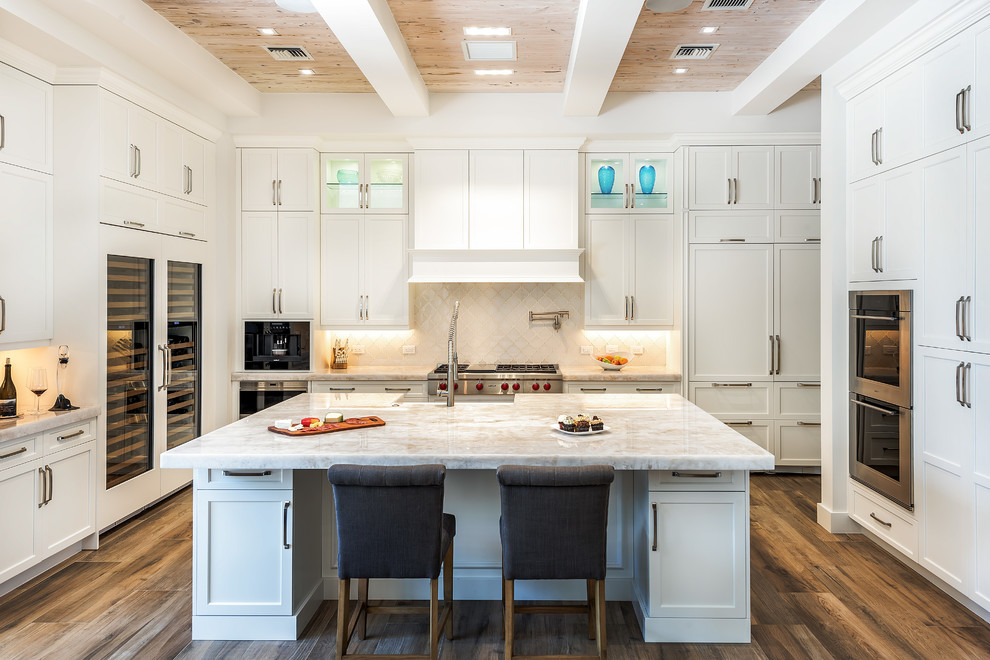 Photo of a mid-sized transitional u-shaped kitchen in Miami with shaker cabinets, white cabinets, white splashback, a farmhouse sink, stainless steel appliances, dark hardwood floors, with island and brown floor.