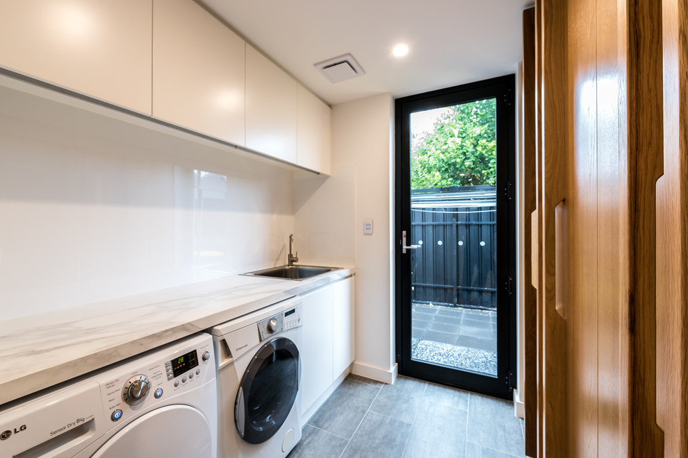 This is an example of a mid-sized contemporary galley dedicated laundry room in Adelaide with a single-bowl sink, flat-panel cabinets, marble benchtops, white walls, ceramic floors, a side-by-side washer and dryer, grey floor, white benchtop and medium wood cabinets.