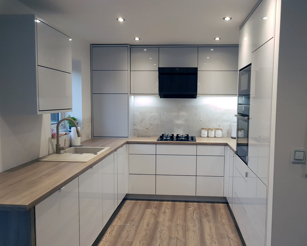 This is an example of a mid-sized modern u-shaped separate kitchen in Other with an integrated sink, flat-panel cabinets, white cabinets, laminate benchtops, black appliances, laminate floors, no island, brown floor and brown benchtop.