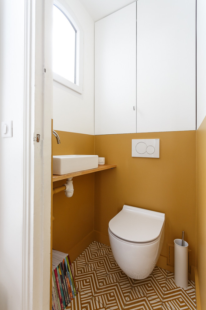 Small powder room in Paris with beaded inset cabinets, white cabinets, a wall-mount toilet, yellow walls, cement tiles, a vessel sink, wood benchtops, yellow floor, beige benchtops and a built-in vanity.