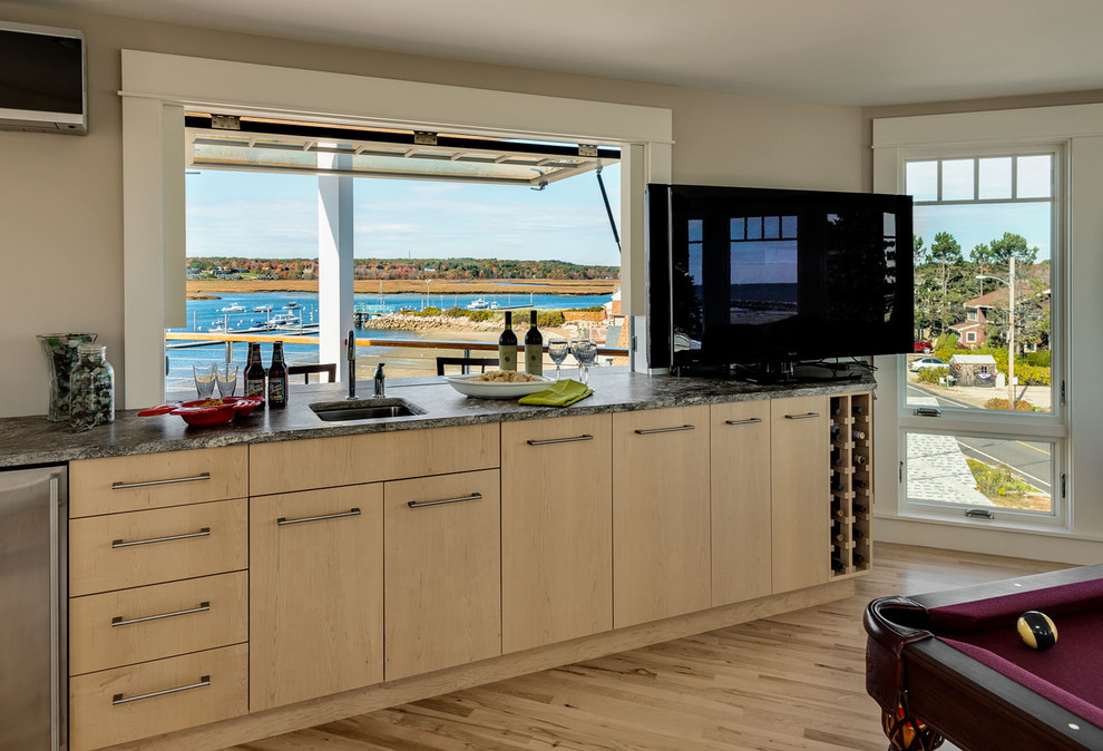 Large beach style l-shaped home bar in Portland Maine with an undermount sink, recessed-panel cabinets, light wood cabinets, granite benchtops, stone slab splashback and medium hardwood floors.