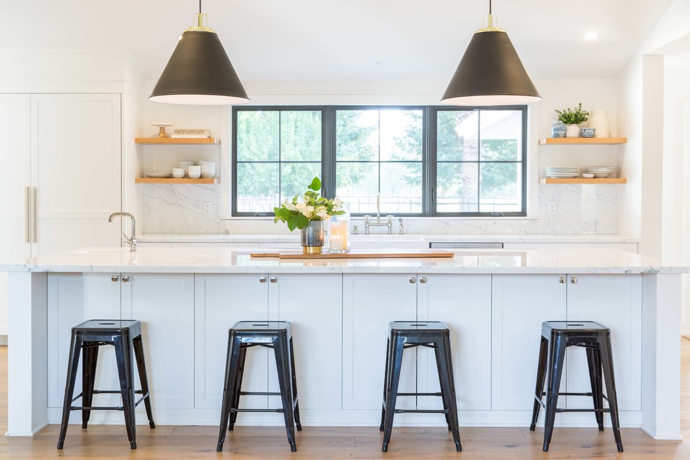 Example of a transitional galley medium tone wood floor and brown floor kitchen design with shaker cabinets, white cabinets, solid surface countertops, marble backsplash, an island, white countertops, a farmhouse sink, white backsplash and paneled appliances