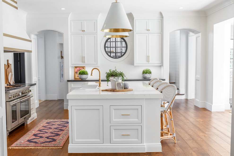 Design ideas for a transitional l-shaped kitchen in Atlanta with a farmhouse sink, recessed-panel cabinets, white cabinets, white splashback, subway tile splashback, stainless steel appliances, dark hardwood floors, with island, brown floor and white benchtop.