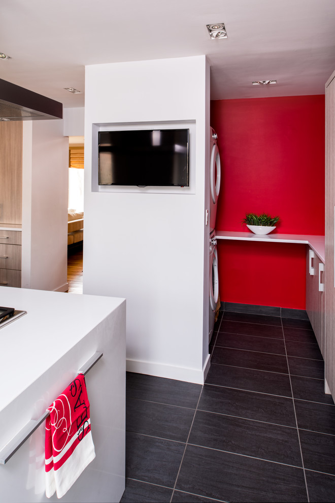 Photo of a small contemporary u-shaped dedicated laundry room in New York with flat-panel cabinets, light wood cabinets, quartz benchtops, red walls, porcelain floors, a stacked washer and dryer and grey floor.