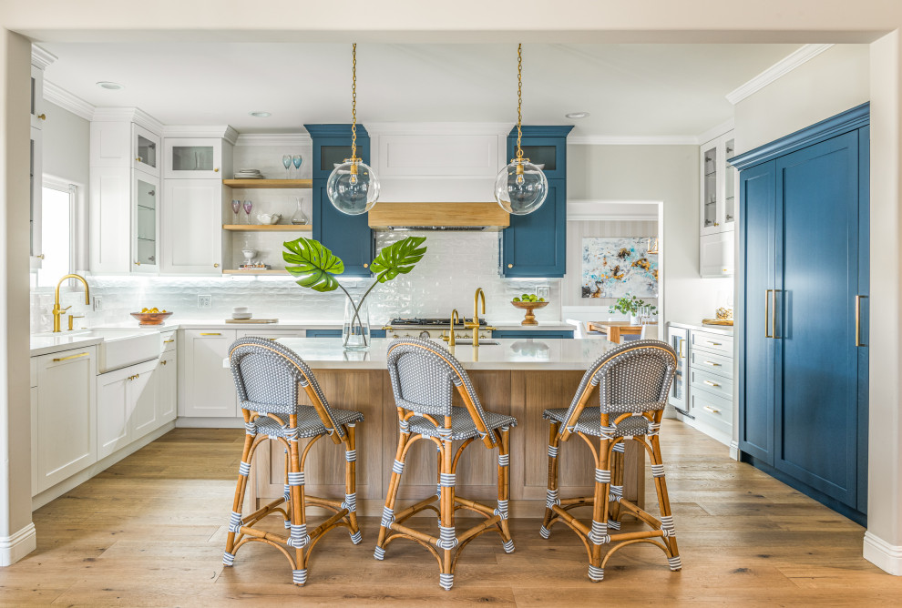 Mid-sized beach style u-shaped open plan kitchen in Los Angeles with a farmhouse sink, shaker cabinets, white cabinets, quartz benchtops, white splashback, subway tile splashback, with island, white benchtop, panelled appliances, medium hardwood floors and brown floor.