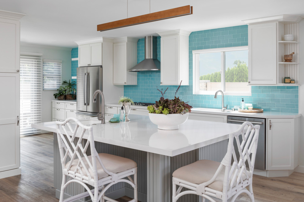 This is an example of a transitional u-shaped kitchen in Orange County with an undermount sink, shaker cabinets, white cabinets, glass benchtops, blue splashback, glass tile splashback, stainless steel appliances, medium hardwood floors, with island, brown floor and white benchtop.