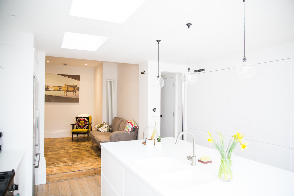 This is an example of a mid-sized modern galley open plan kitchen in London with an integrated sink, flat-panel cabinets, white cabinets, white splashback, stainless steel appliances, light hardwood floors, with island, brown floor and white benchtop.