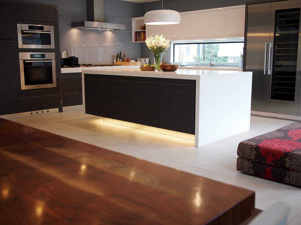 This is an example of a mid-sized contemporary l-shaped eat-in kitchen in Los Angeles with a double-bowl sink, flat-panel cabinets, stainless steel appliances, with island, white floor, dark wood cabinets, quartz benchtops, grey splashback, glass sheet splashback, porcelain floors and white benchtop.