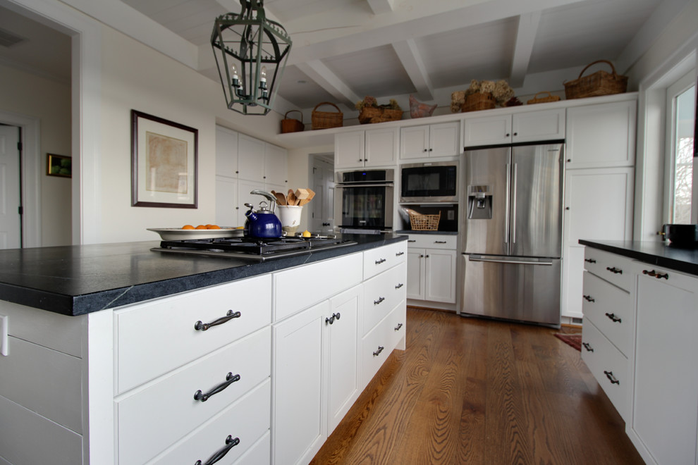Photo of a mid-sized transitional l-shaped separate kitchen in Other with a farmhouse sink, flat-panel cabinets, white cabinets, quartz benchtops, stainless steel appliances, medium hardwood floors, with island, brown floor and black benchtop.