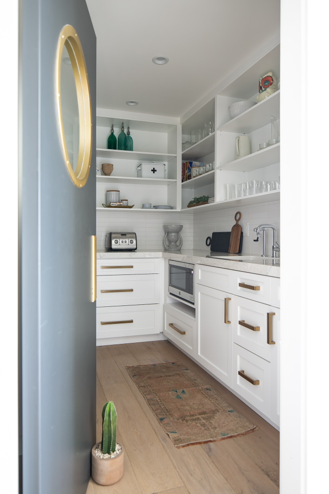 Inspiration for a beach style kitchen pantry in Orange County with an undermount sink, shaker cabinets, white cabinets, white splashback, stainless steel appliances, light hardwood floors, no island, beige floor and white benchtop.