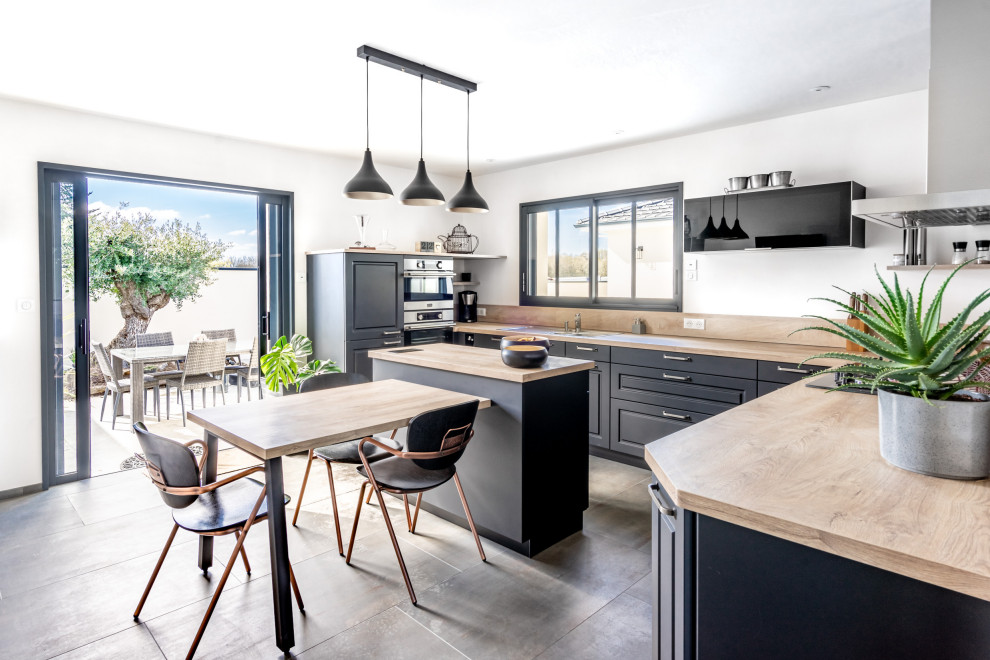 This is an example of a large country u-shaped open plan kitchen in Nantes with an undermount sink, beaded inset cabinets, black cabinets, wood benchtops, beige splashback, timber splashback, panelled appliances, ceramic floors, with island, beige floor and beige benchtop.