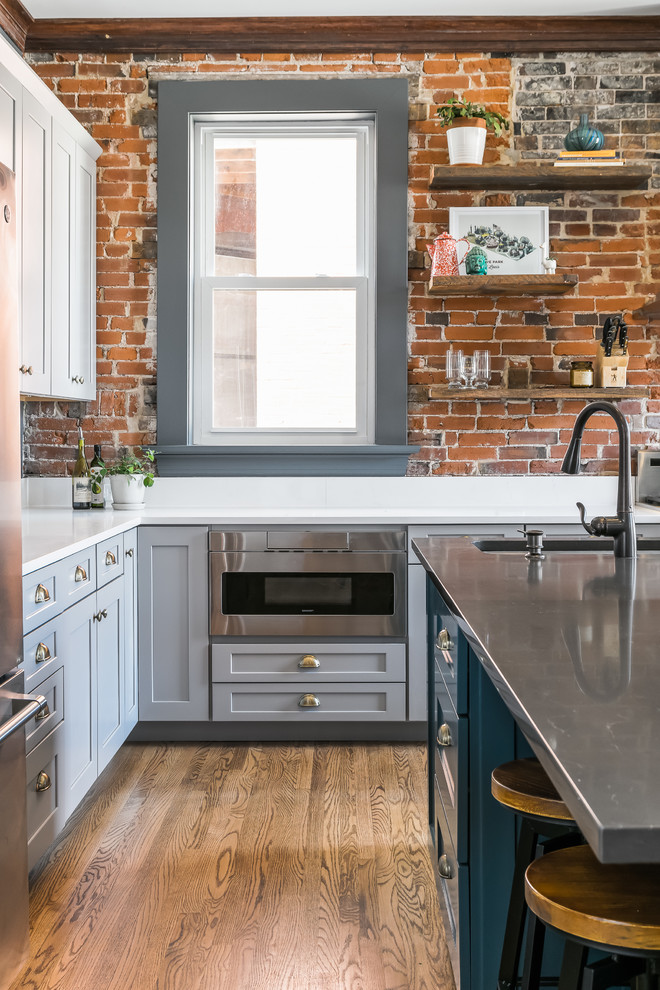 Photo of a mid-sized industrial kitchen in St Louis with a single-bowl sink, shaker cabinets, grey cabinets, quartz benchtops, brick splashback, stainless steel appliances, medium hardwood floors, with island and white benchtop.