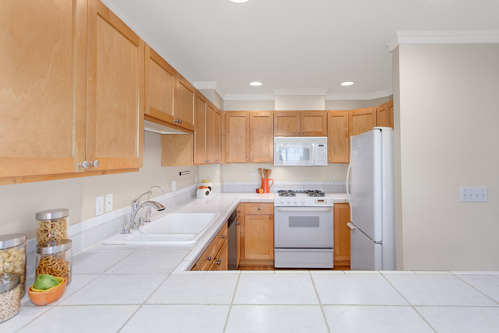 This is an example of a mid-sized transitional u-shaped eat-in kitchen in Seattle with a double-bowl sink, shaker cabinets, light wood cabinets, tile benchtops, white appliances, light hardwood floors and no island.