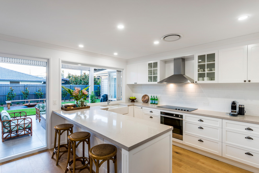 Design ideas for a mid-sized beach style u-shaped eat-in kitchen in Gold Coast - Tweed with a farmhouse sink, shaker cabinets, white cabinets, quartz benchtops, white splashback, ceramic splashback, stainless steel appliances, light hardwood floors, with island and brown floor.