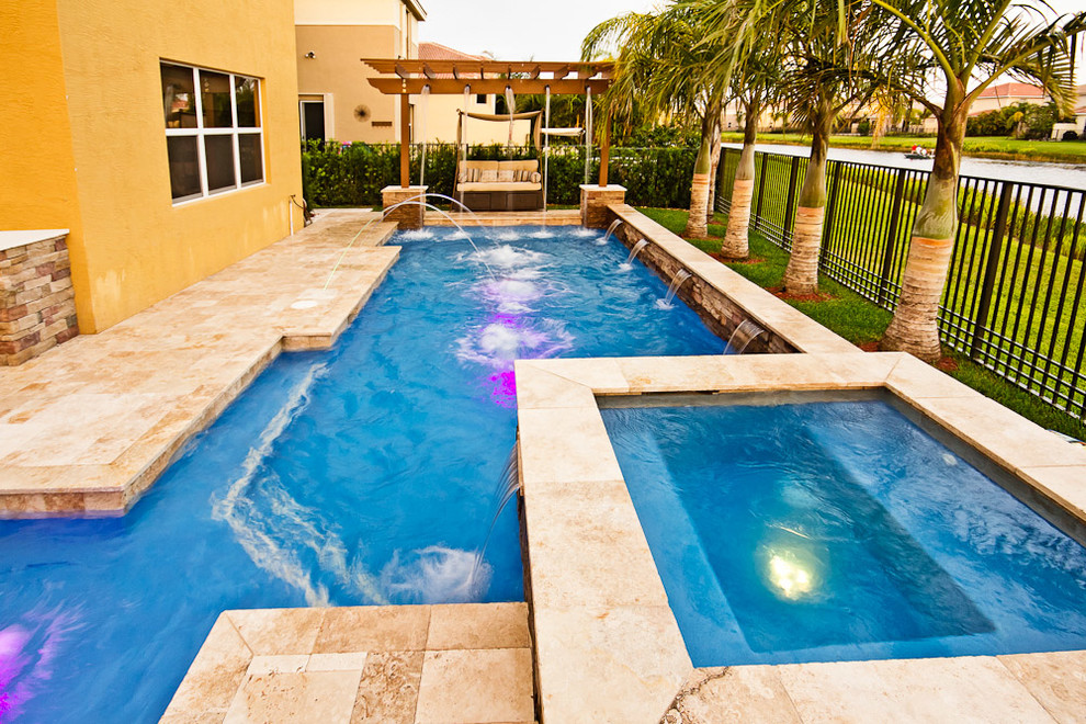 This is an example of a large mediterranean backyard custom-shaped lap pool in Miami with a water feature and natural stone pavers.