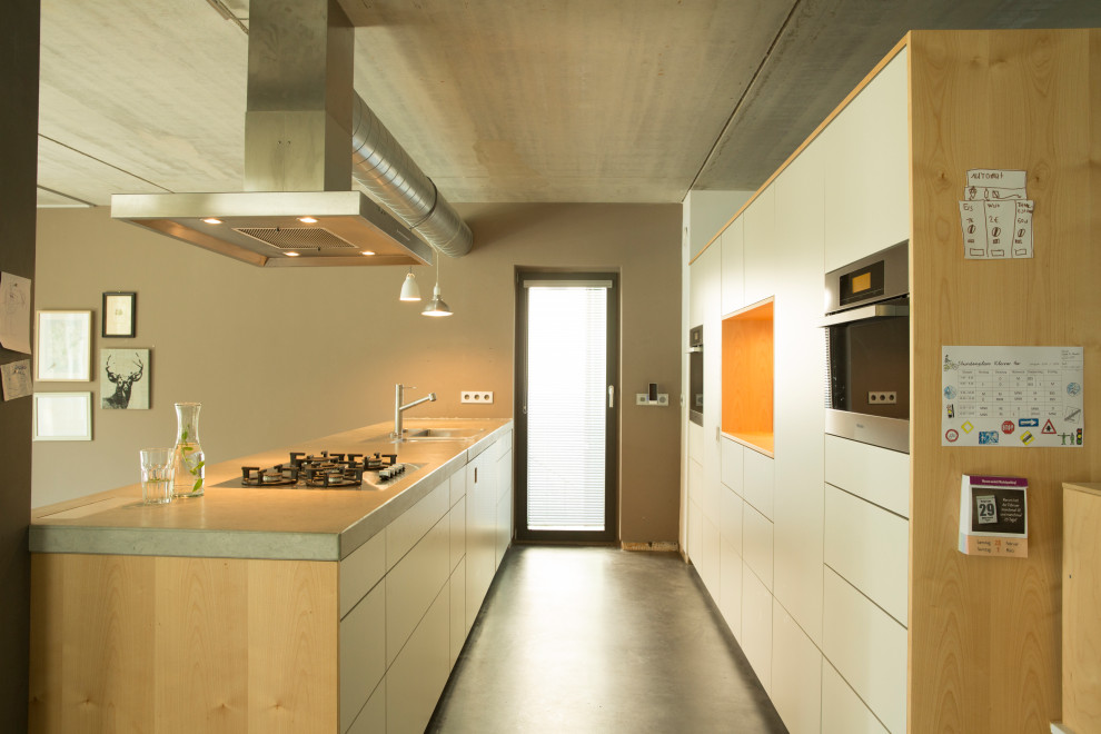 Large industrial galley eat-in kitchen in Stuttgart with a drop-in sink, flat-panel cabinets, white cabinets, limestone benchtops, concrete floors, a peninsula, black floor and grey benchtop.