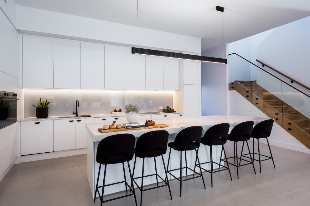 Photo of a contemporary l-shaped kitchen in Adelaide with flat-panel cabinets, white cabinets, grey splashback, black appliances, with island, grey floor and white benchtop.