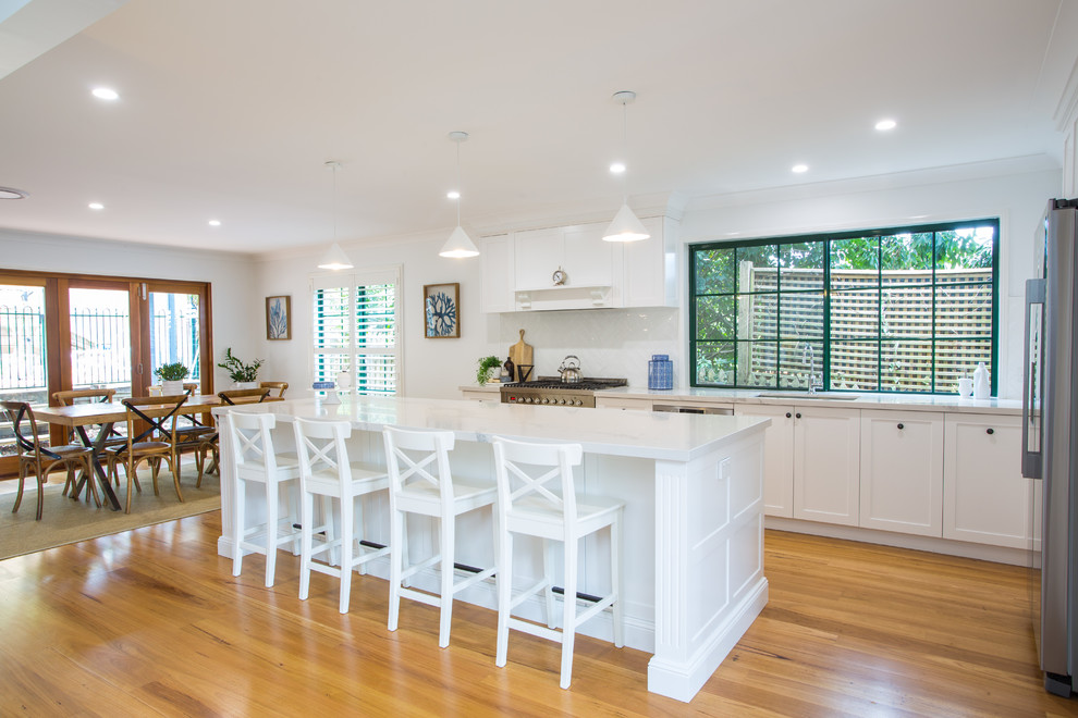 Expansive traditional galley eat-in kitchen in Brisbane with a single-bowl sink, shaker cabinets, white cabinets, solid surface benchtops, white splashback, subway tile splashback, stainless steel appliances, medium hardwood floors, with island, brown floor and white benchtop.